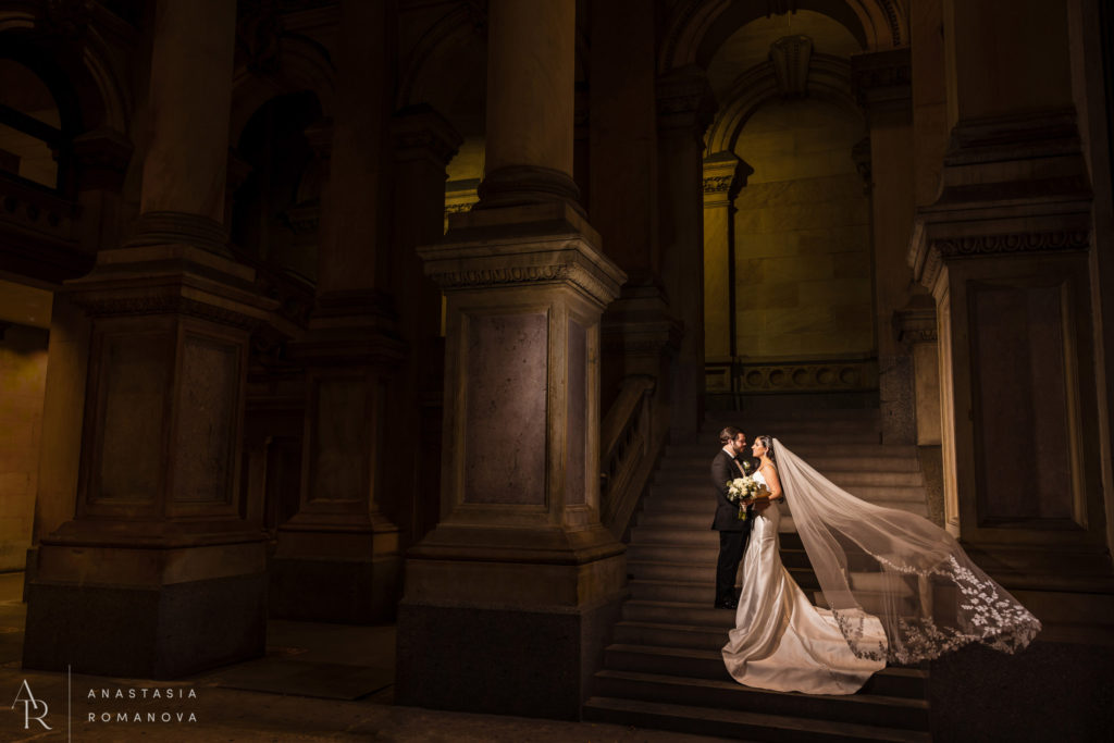 Bride and groom standing on the steps of Philadelphia City Hall with a cathedral veil flowing in the air in dramatic light