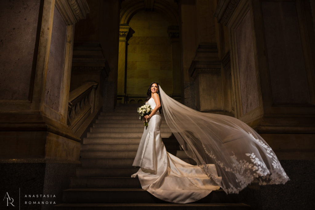 Bride with long curly hair on the steps of the City Hall with a long cathedral veil flowing in the air in beautiful light